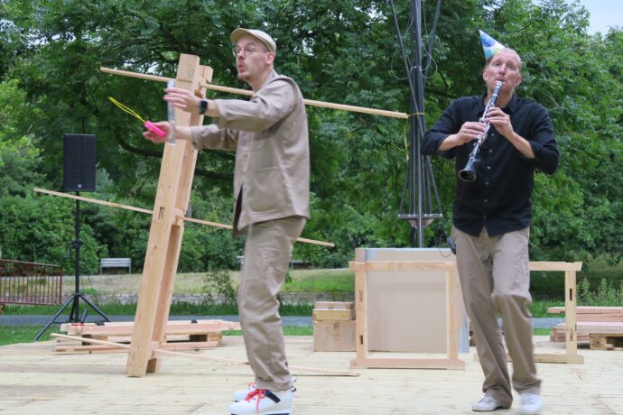 Pour toi Erik - buitenscene met een man die fluit speelt en een man die een houtbouw in elkaar probeert te zetten